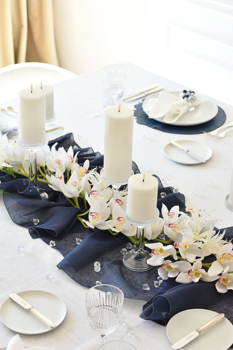 Elegant table setting with candles, white plates, and blue napkins on a white tablecloth.