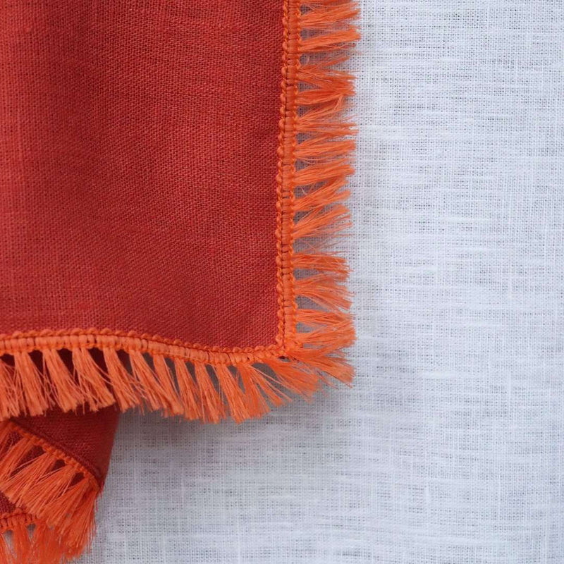 Close-up of the fringe detailing on the orange linen napkin on a white tablecloth.