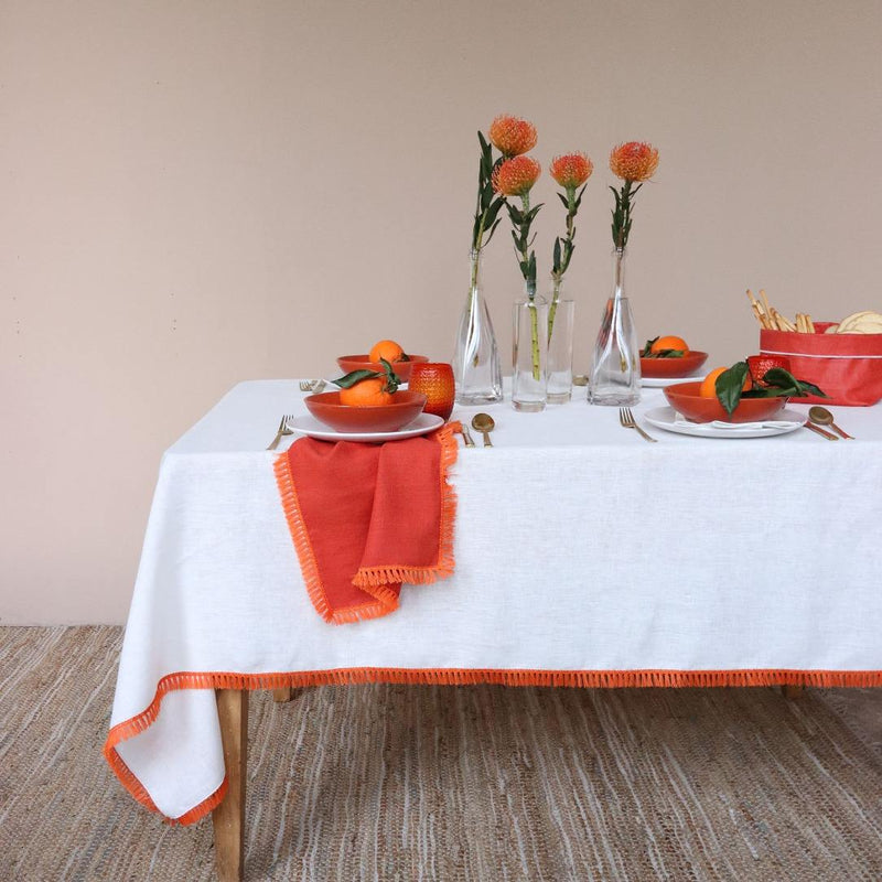 Orange linen napkin with fringe on the side of the table underneath a bowl of oranges.