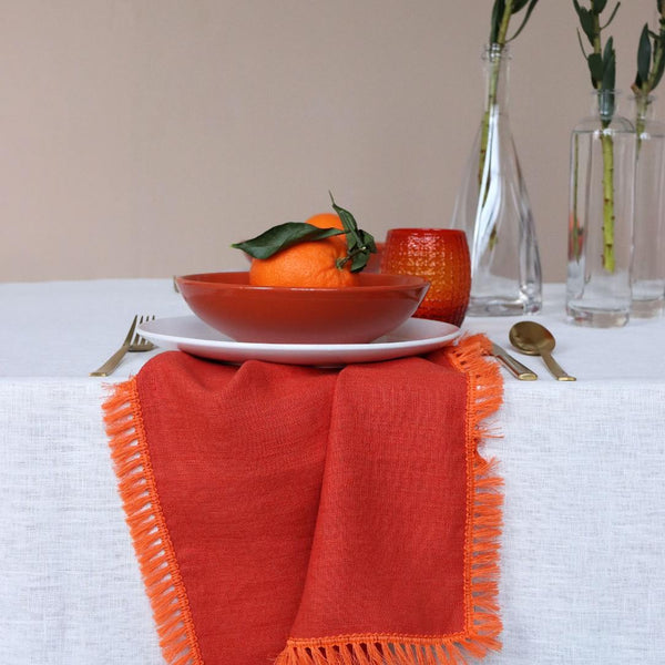 Orange linen napkin with fringe by Giardino Segreto underneath a bowl of oranges.