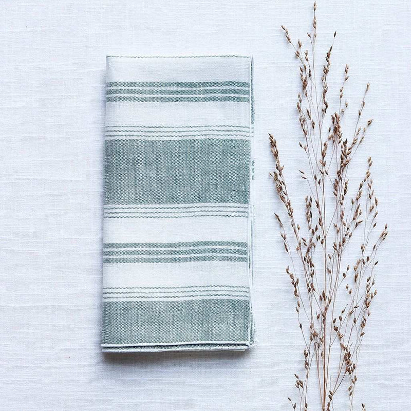 Folded Riva green and white striped linen napkin with piped edge on white tablecloth.