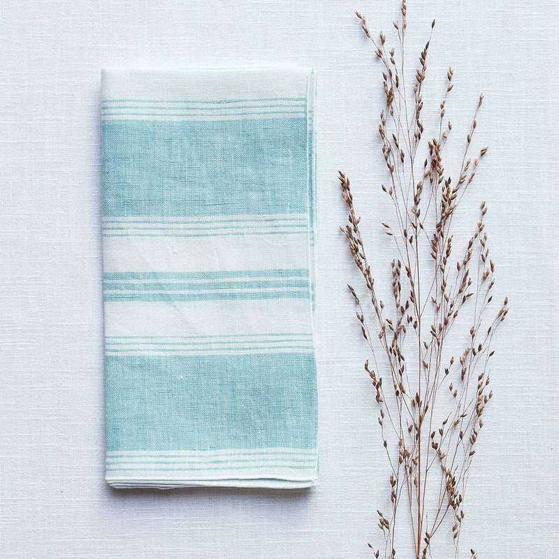 Folded Riva turquoise and white striped linen napkin with piped edge on white tablecloth 
