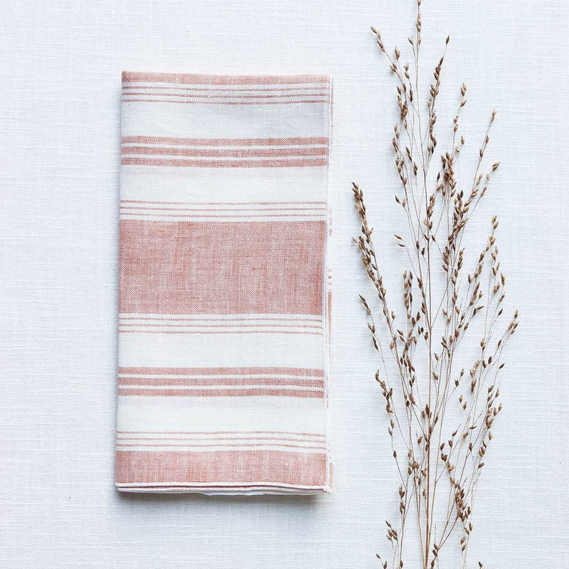 Linen napkin with coral and white irregular stripes folded on white tablecloth, sold at Amiramour.
