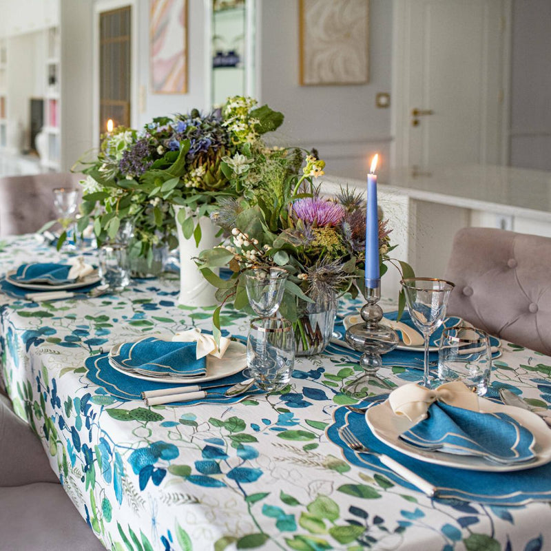 Blue Petali napkins with wave borders and ecru embroidery along the borders styled on table setting with green plants, sold at Amiramour.