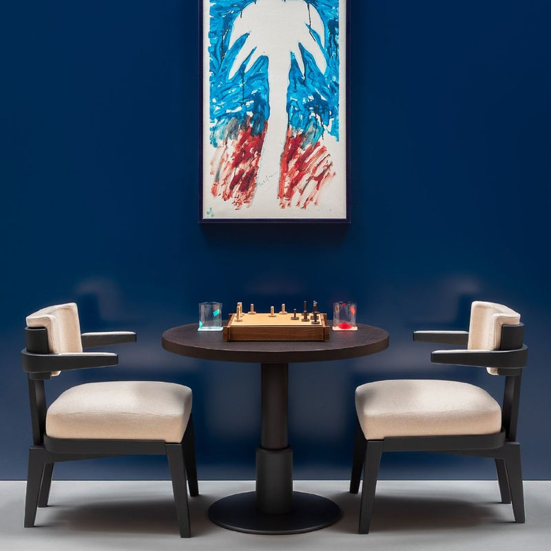 Round table with Open Structura Triple Game Box by Giobagnara and chairs against a blue wall with abstract art