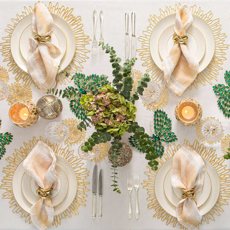 Elegant table setting with gold and white plates, napkins, and greenery on a white tablecloth.