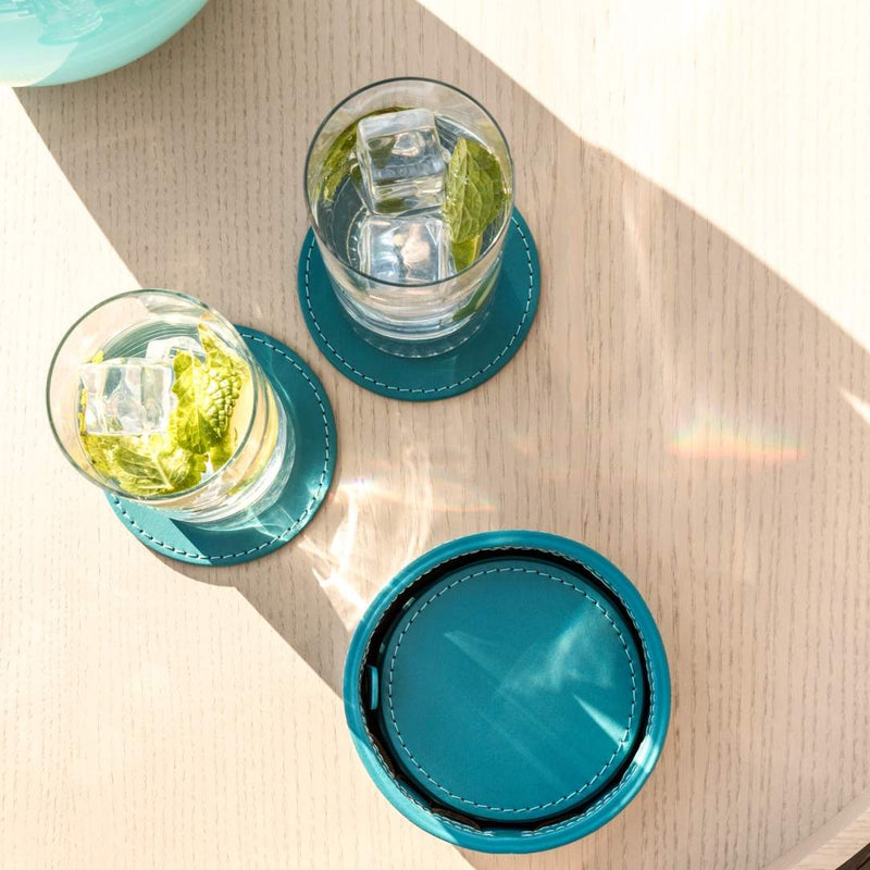 Two glasses of water with ice and a green leaf on turquoise coasters on a light wooden surface.