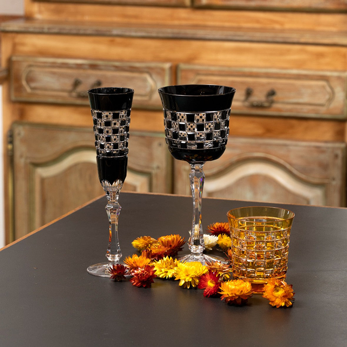 Two decorative wine glasses Black Diamond with a patterned top on a table with flowers and a glass of yellow liquid.