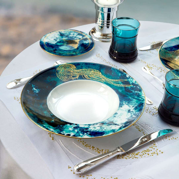 Table setting with blue and gold Ocean Bleu by Haviland plates, glasses, and cutlery on a white tablecloth.