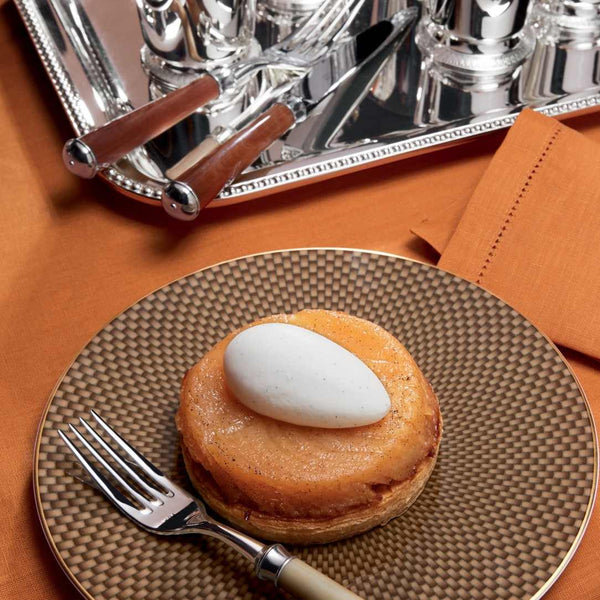 Small dessert with a white topping on a textured plate Raynaud Trésor brown porcelain bread plate with gold-accented honeycomb design with a fork, set against an orange background.
