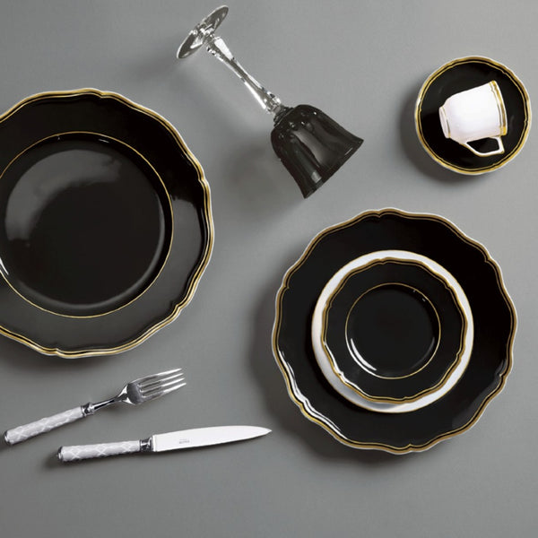 Black dinnerware set featuring luxury porcelain small plate mazurka gold and black raynaud with scalloped contour and gold edging on a gray surface, including plates, a knife, fork, and glass.