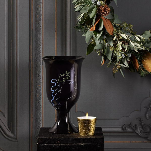 Black decorative vase with a gold candle on a dark surface, with a wreath in the background.
