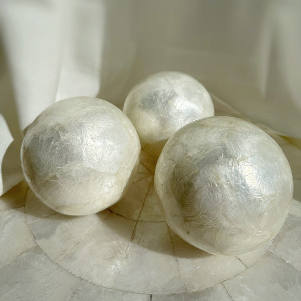Three round ivory-colored capiz shell decorative balls are displayed on a capiz placemat. Each ball has a subtle, natural pattern and a glossy finish.
