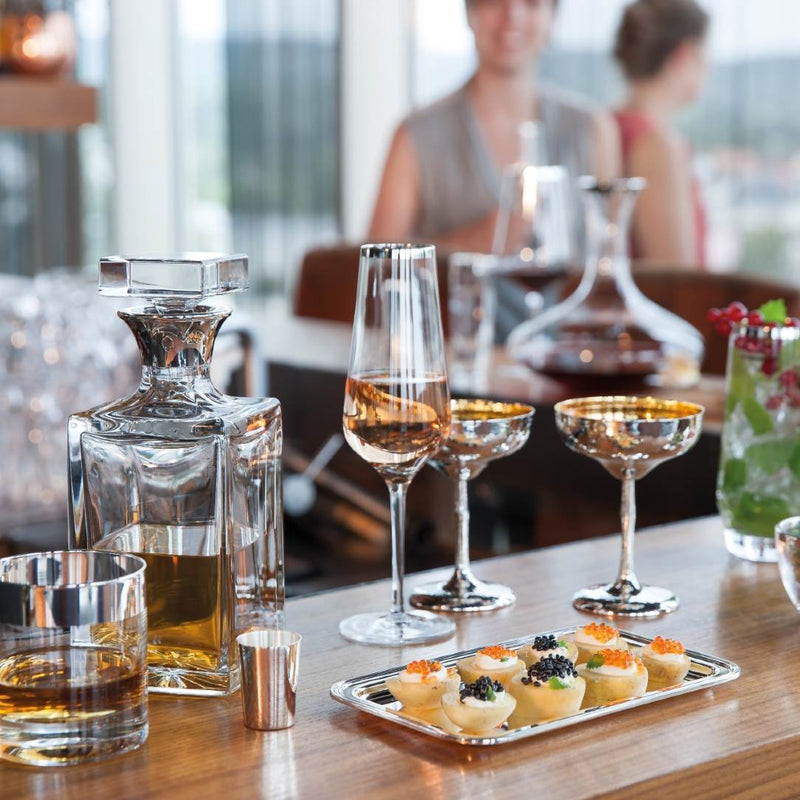 Glass decanter, Elegant Crystal Champagne Flute with Hand-Applied Silver Rim – Avantgarde by Sonja Quandt, and appetizers on a table with blurred people in the background