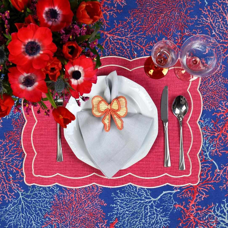 Decorative table setting with red flowers, a red placemat, white napkin with a bow, and cutlery on a blue and red patterned tablecloth.