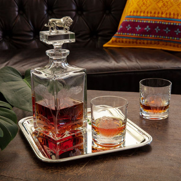Crystal decanter with lion stopper on a tray with two glasses of whiskey, set against a dark leather couch.