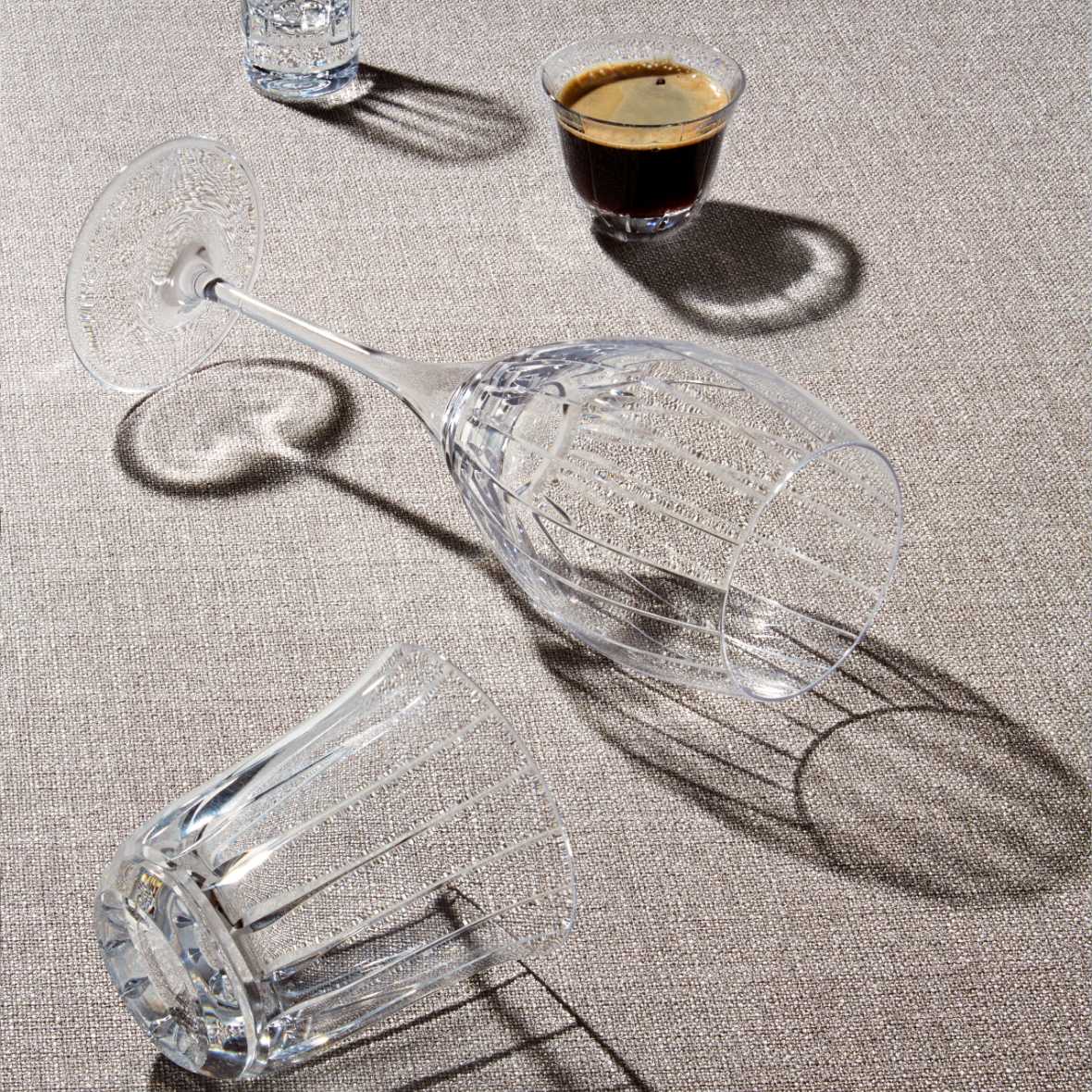 Hand-cut crystal water glass with fine bevels from the Saint-Rémi collection by Cristallerie de Montbronn on a textured surface with a small glass of dark liquid nearby.