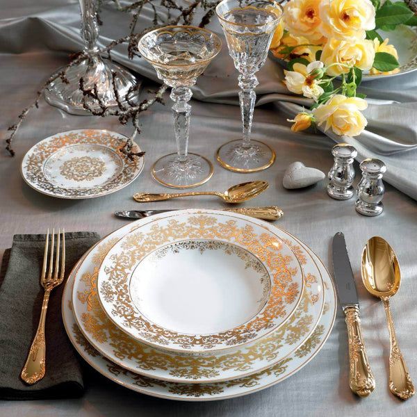 Elegant table setting with gold and white plates, glasses, and silverware on a gray tablecloth.