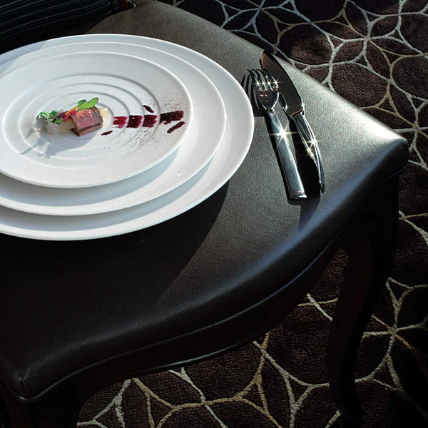 White plate with a small dish of food on a black chair with a patterned carpet.