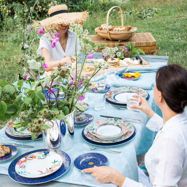 Two women setting a table outdoors with floral decorations and colorful plates.