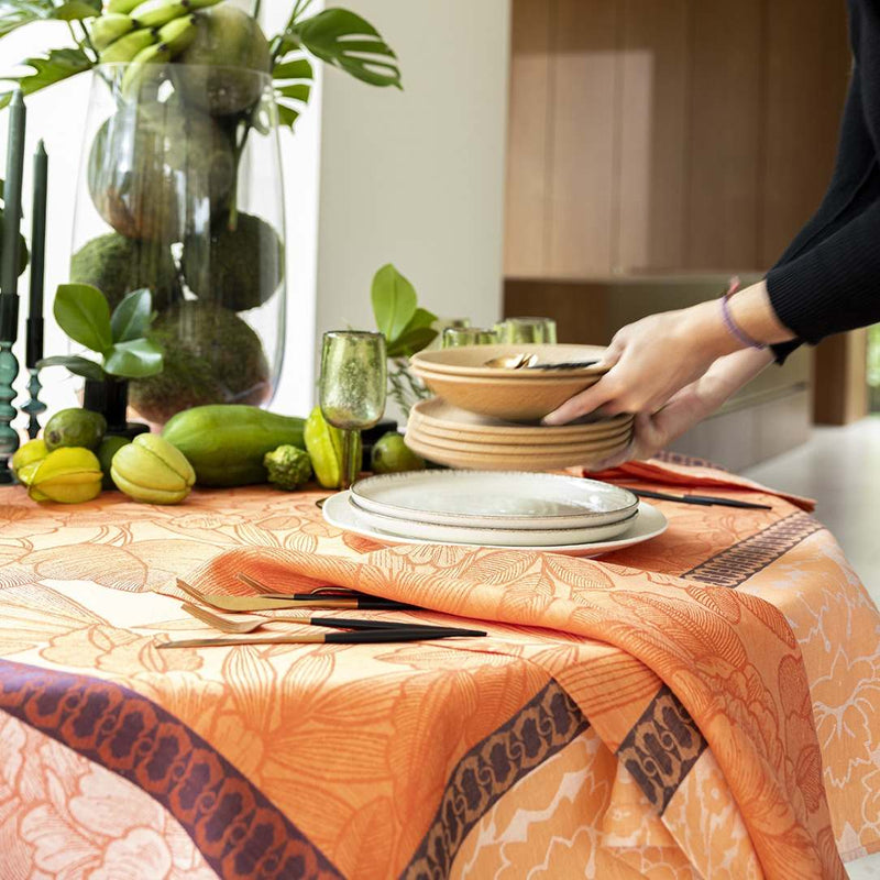 Person setting a table with plates and cutlery, surrounded by greenery and a vase.