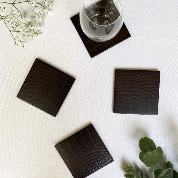 Set of four dark brown crocodile-patterned coasters on a light surface with a glass of water and greenery.
