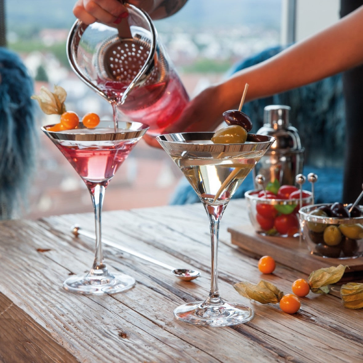 Cocktail being poured into two handcrafted Wine Glasses Crystal with Silver Rim – Sonja Quandt on a wooden table with a scenic background