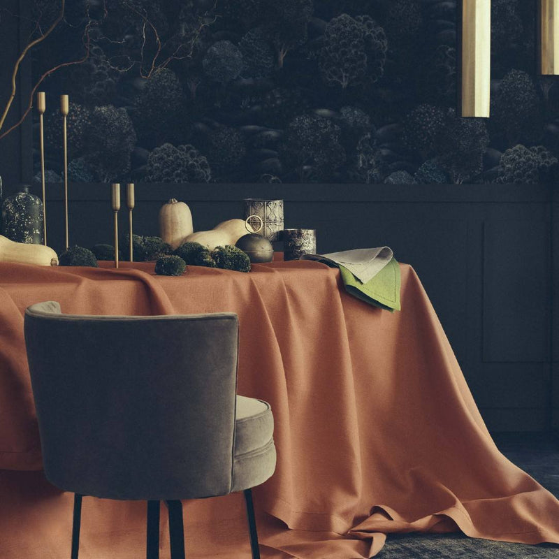 Dining table with a rust-colored tablecloth and gray chair against a dark wall with nature-themed wallpaper.