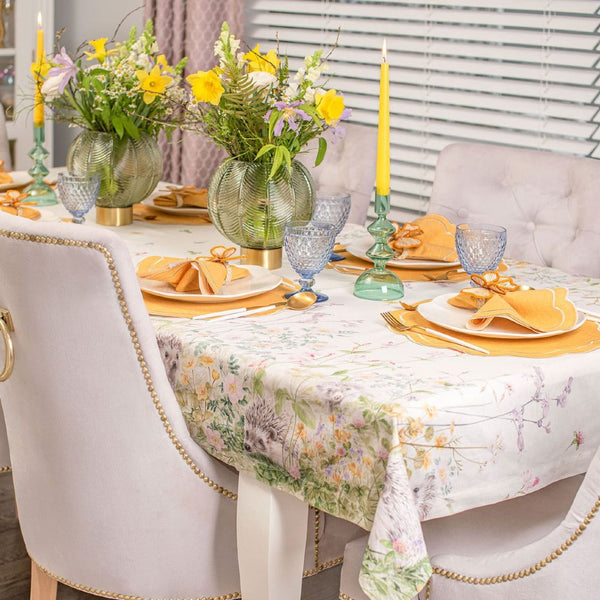 A beige cotton tablecloth with a floral and animal pattern, draped over a table with a yellow tablecloth on the side.