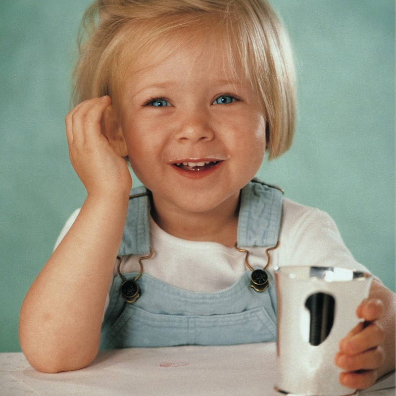 Child smilling holding a silver-plated baby cup with minimalist design by Ercuis against a light green background