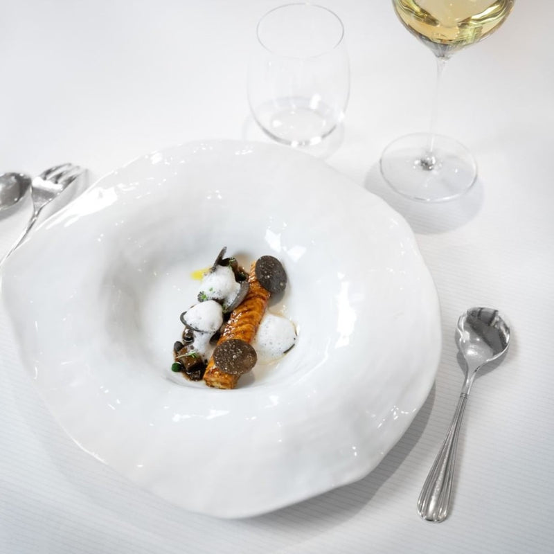 Delicate dish on a Set of 2 large deep dinner plates white No7 Indulge by Sarah-Linda porcelain with two wine glasses and cutlery on a white tablecloth.