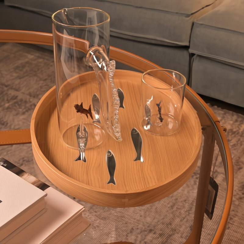 Clear glass pitcher with fish design on a wooden tray on a coffee table.