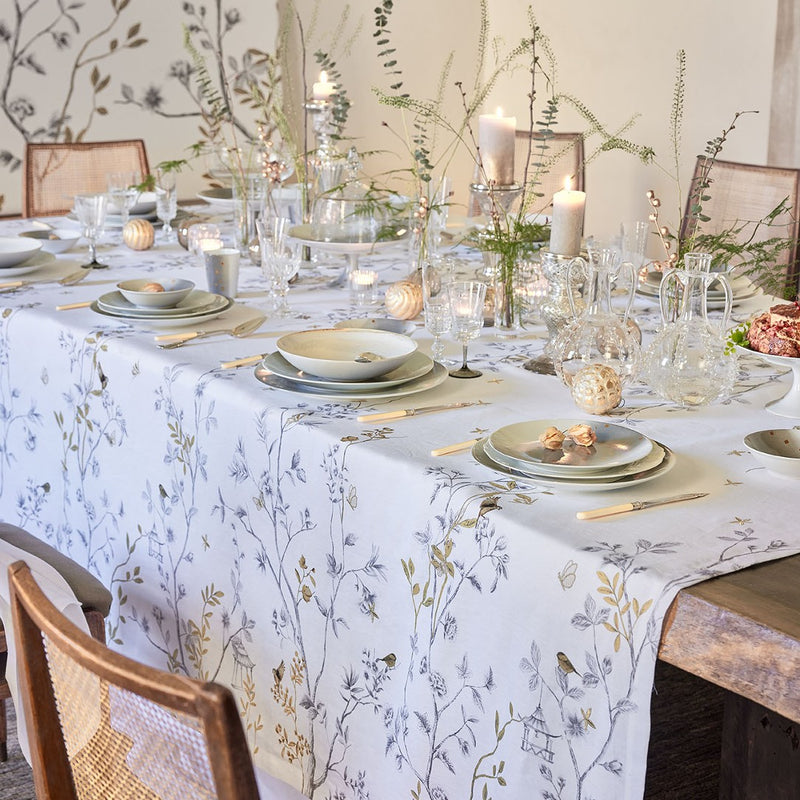 White linen tablecloth with bird and trees embroidery on a table with plates, glassware and candles