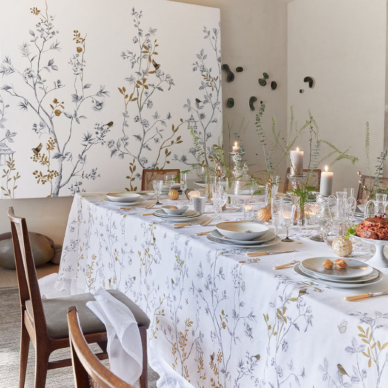 White linen tablecloth with bird and trees embroidery on a table with plates, glassware and candles and a big tree painting on the background