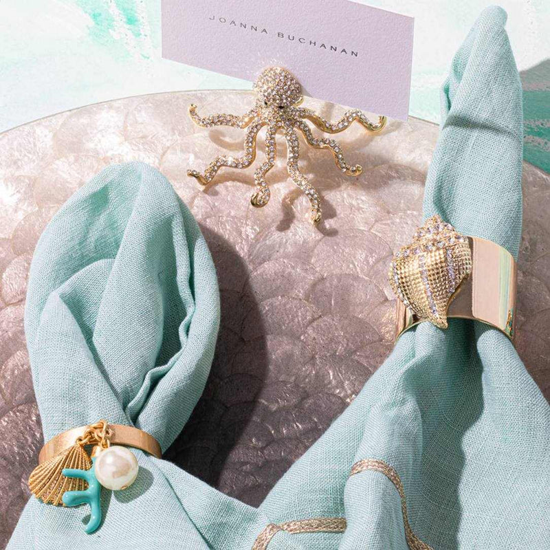 Decorative napkin rings with shell and and Gold octopus place card holder with crystal accents by Joanna Buchanan on a textured surface.