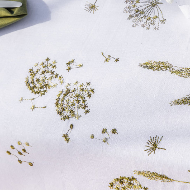 Close-up of a white linen tablecloth with gold embroidery featuring ear of wheat and country plants, displayed on a table.