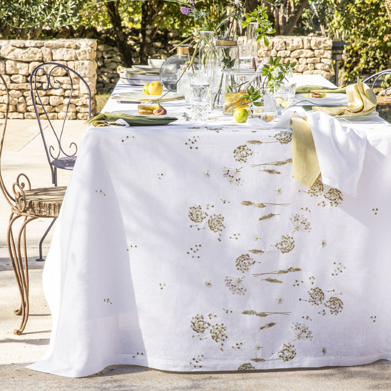 A white linen tablecloth with gold embroidery featuring ear of wheat and country plants, displayed on a table with tableware and vases of flowers