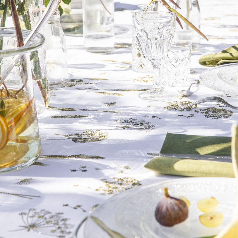 A white linen tablecloth with gold embroidery featuring ear of wheat and country plants, displayed on a table with glassware and napkin.