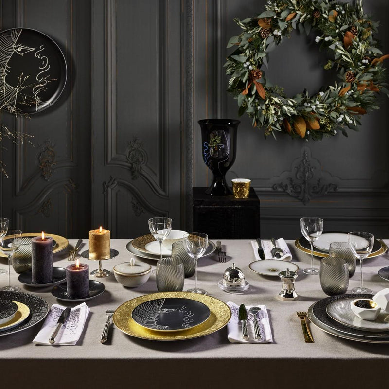 Elegant dining table setting with gold and black plates, candles, and a wreath on a dark wall.