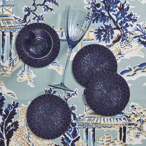 Set of four dark blue beaded coasters on a patterned tablecloth with a glass and bowl.
