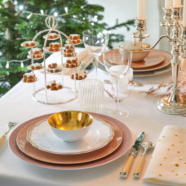 Elegant table setting with gold and pink plates, glasses, and a Christmas tree in the background.