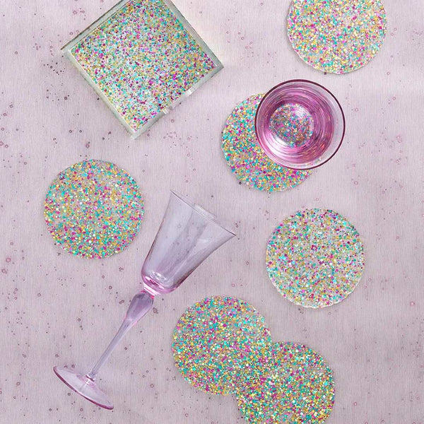 Glittery coasters with a glass of pink liquid and a purple glass on a textured surface.