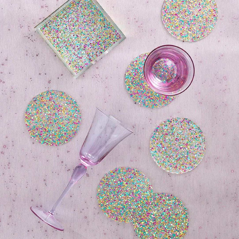Glittery coasters with a glass of pink liquid and a purple glass on a textured surface.