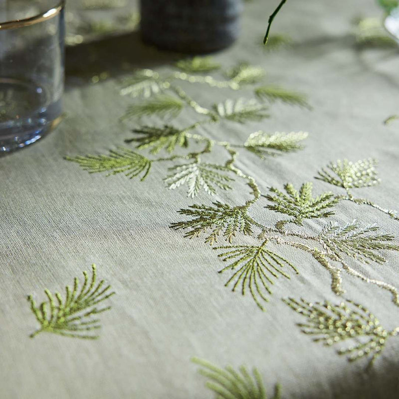 Embroidered green leaves on a beige fabric with a glass and plant in the background.