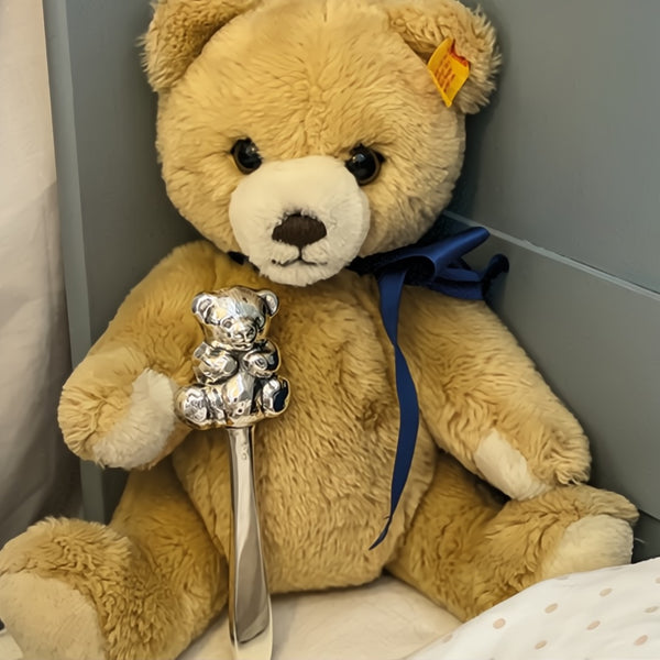 Beige teddy bear holding a silver teddy bear figurine with a blue ribbon.