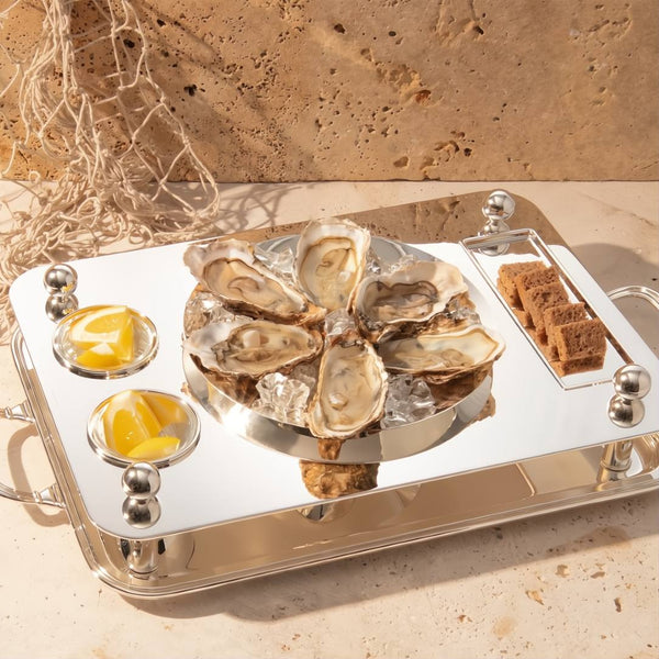 Rectangular silver-plated oyster set with ice bowl and compartments by Sonja Quandt with oysters on a bed of ice, lemon wedges, and bread on a stone surface.