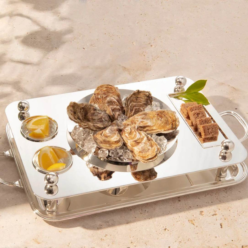 Rectangular silver-plated oyster set with 23 cm bowl by Sonja Quandt with oysters on ice, lemon wedges, and brown crackers on a beige background