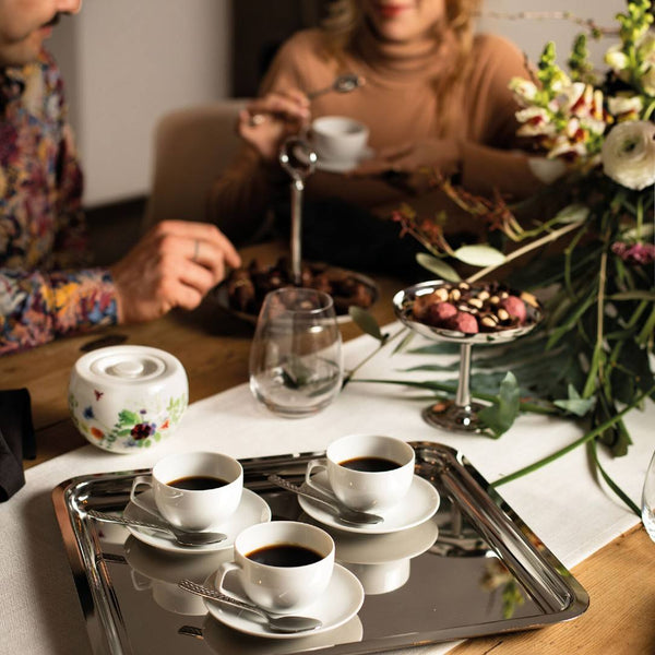 Two people sitting at a table with coffee cups, a tray of snacks, and a floral arrangement.