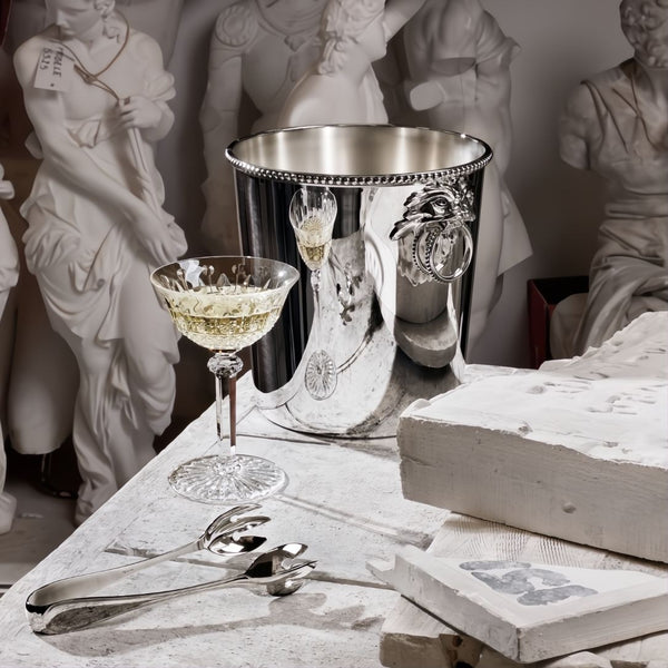 Decorative table setting with a silver ice bucket, glass, and Régency silver-plated ice tongs by Ercuis against classical statue background