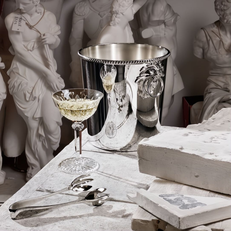 Decorative table setting with a silver ice bucket, glass, and Régency silver-plated ice tongs by Ercuis against classical statue background